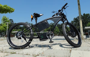 A black e-bike parked on tiles outdoors in the sunlight. Trees can be seen in the background.